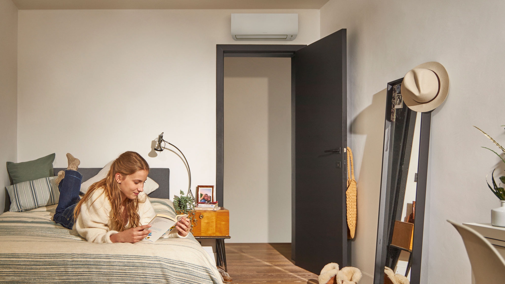 Žena si čte na posteli v útulné ložnici s klimatizačním zařízením s tepelným čerpadlem namontovaným nad dveřmi, které zajišťuje energeticky účinnou regulaci klimatu.