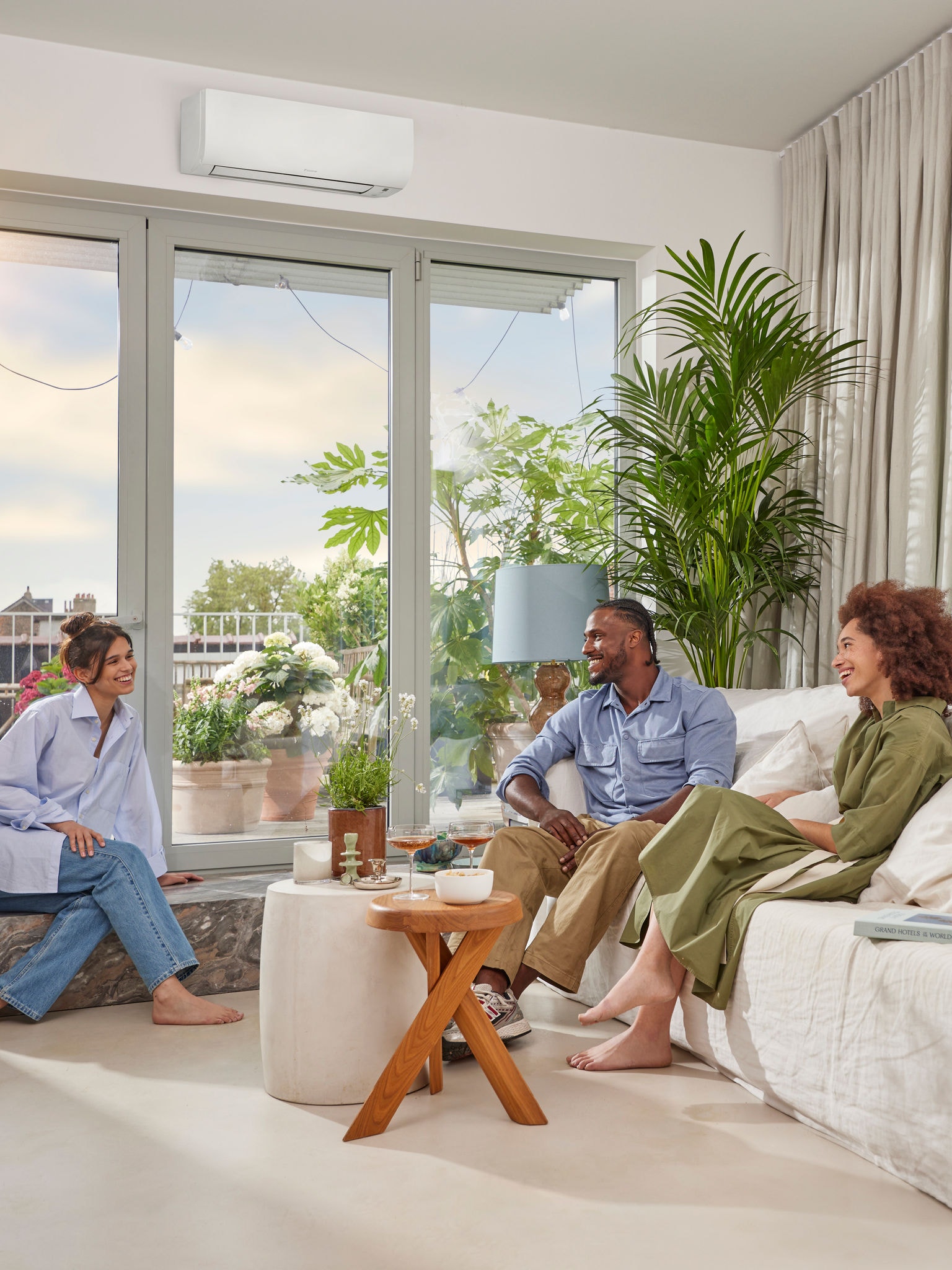 Muž a dvě ženy sedí a povídají si ve světlé, vzdušné místnosti s bílými stěnami a nenápadným bílým tepelným čerpadlem namontovaným na zdi nad velkým oknem za slunečného dne