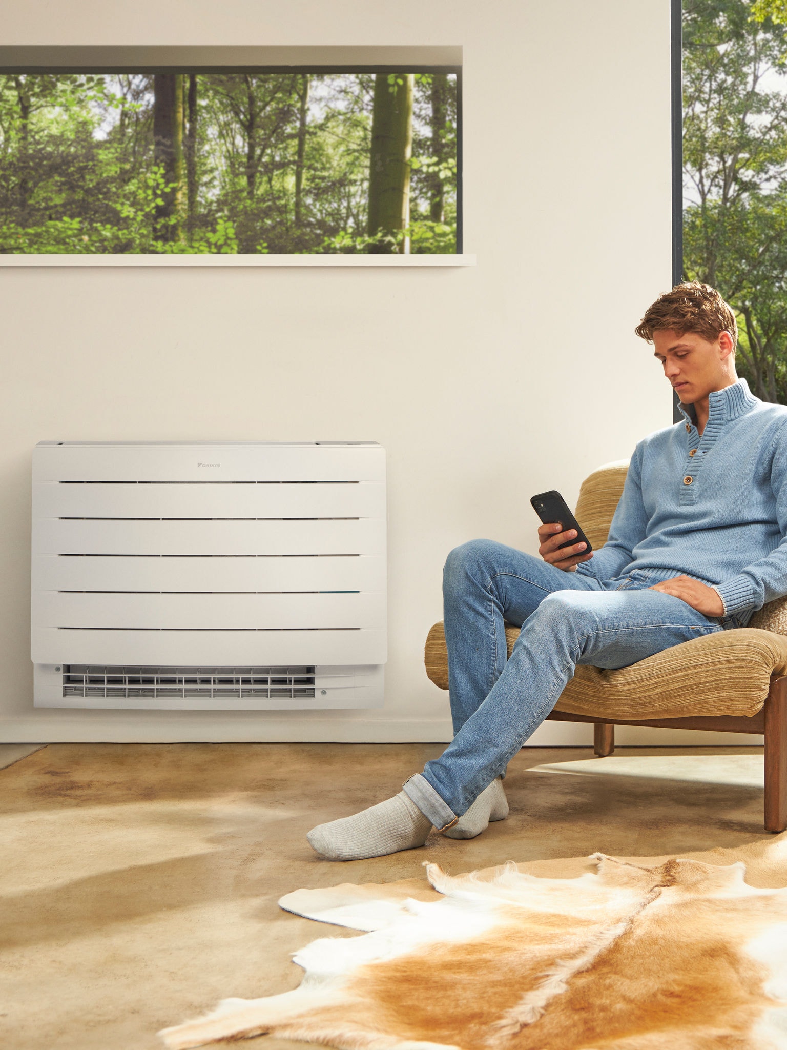 Muž sedící ve světlé místnosti a dívající se na svůj telefon s tepelným čerpadlem na zdi za sebou