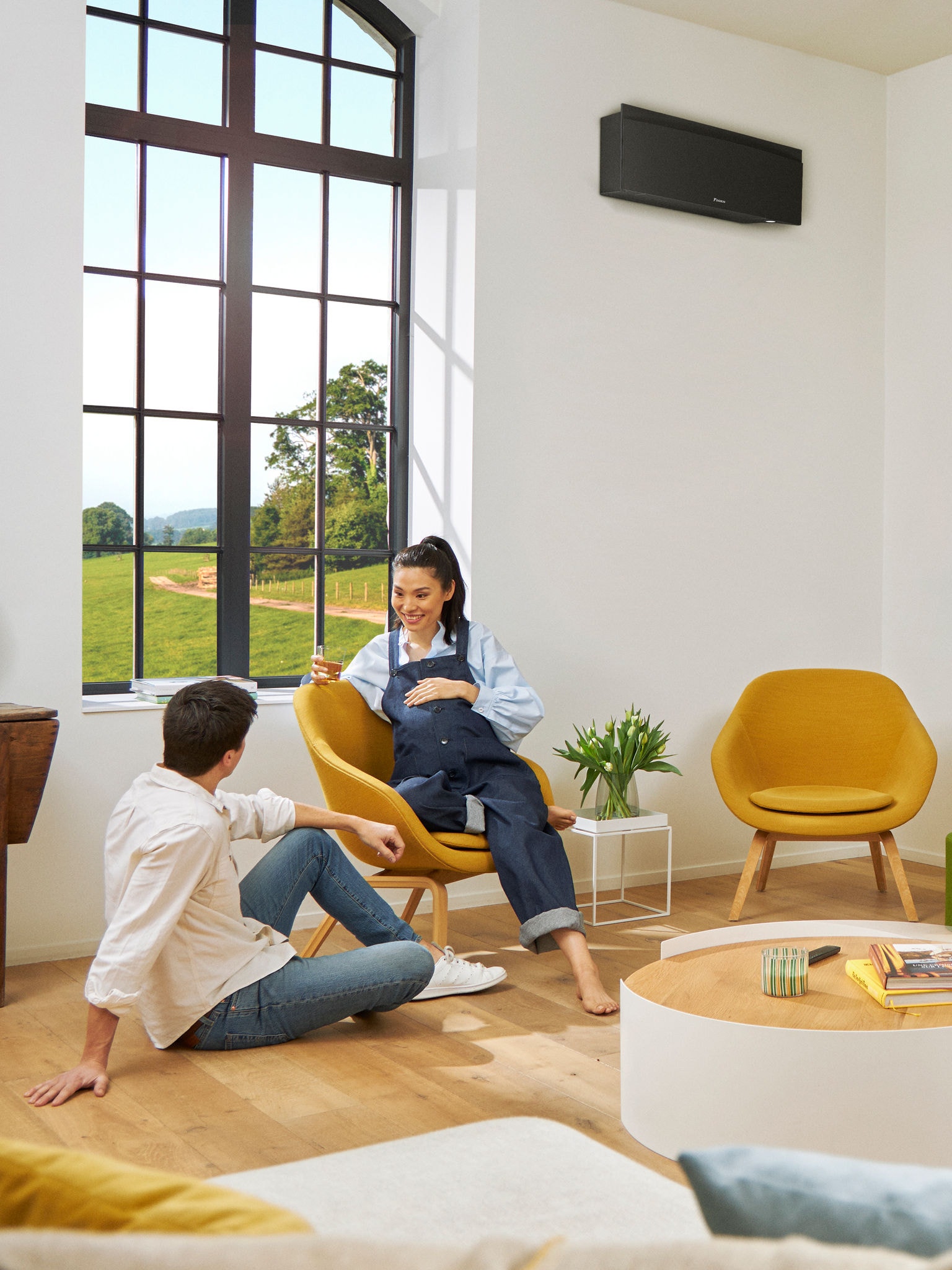 Muž a žena sedící před velkým oknem ve světlé místnosti s černým nástěnným tepelným čerpadlem na zdi za nimi