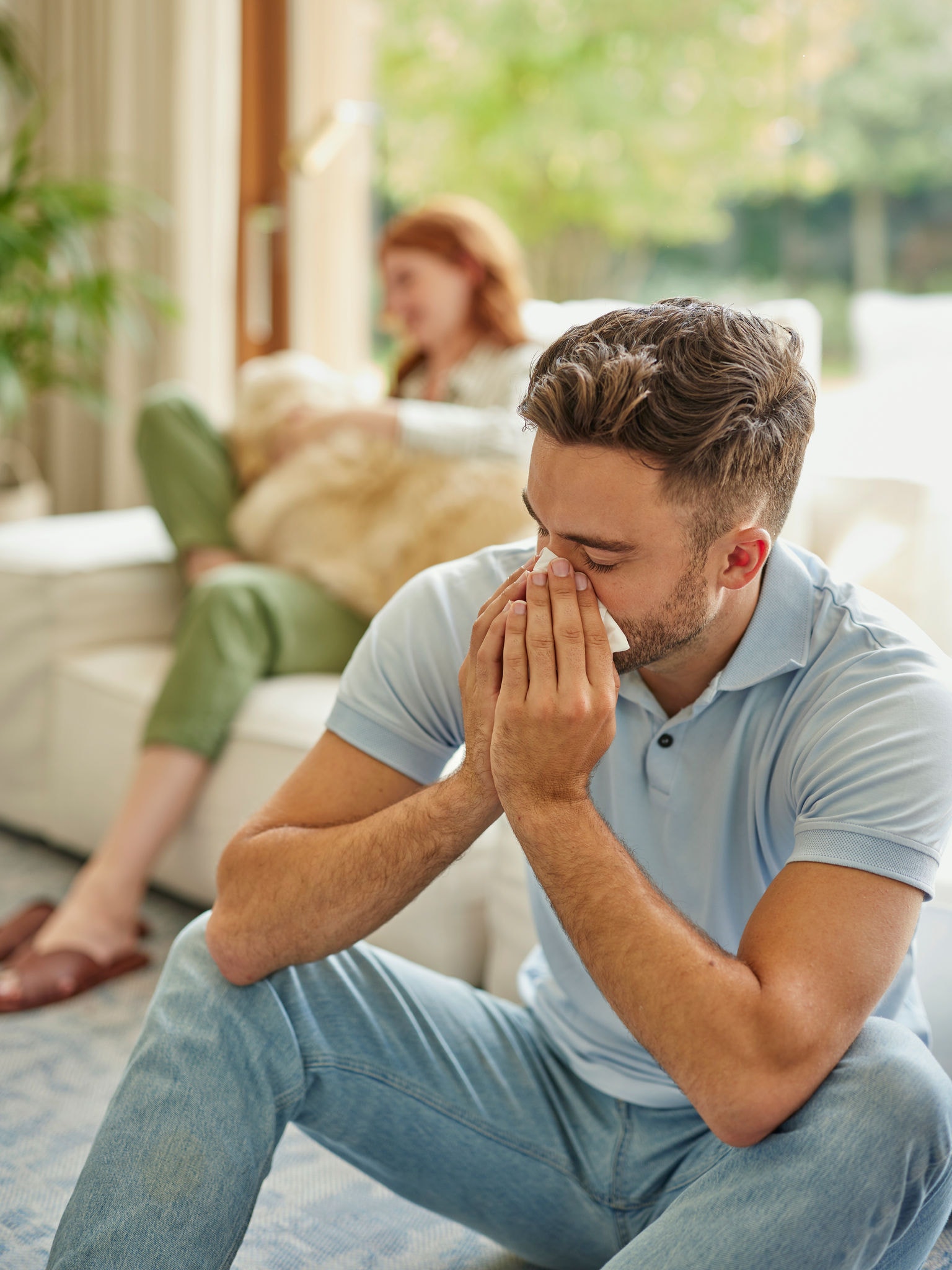 Žena v pozadí na bytovém doplňku z textilu se zlatým retrívrem na klíně, muž v popředí kýchá