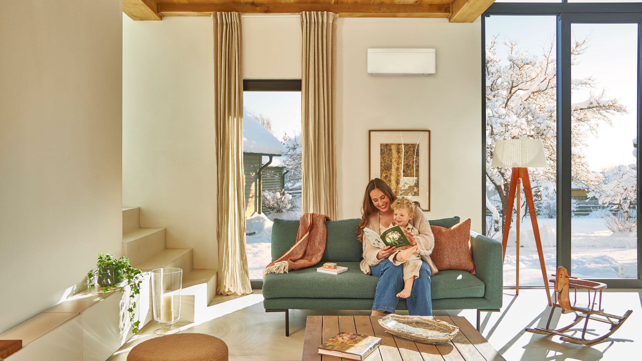 Matka sedí na gauči pod nástěnným tepelným čerpadlem vzduch-vzduch a čte si dětskou knížku s batoletem na klíně a užívá si tepla, zatímco venku sněží.